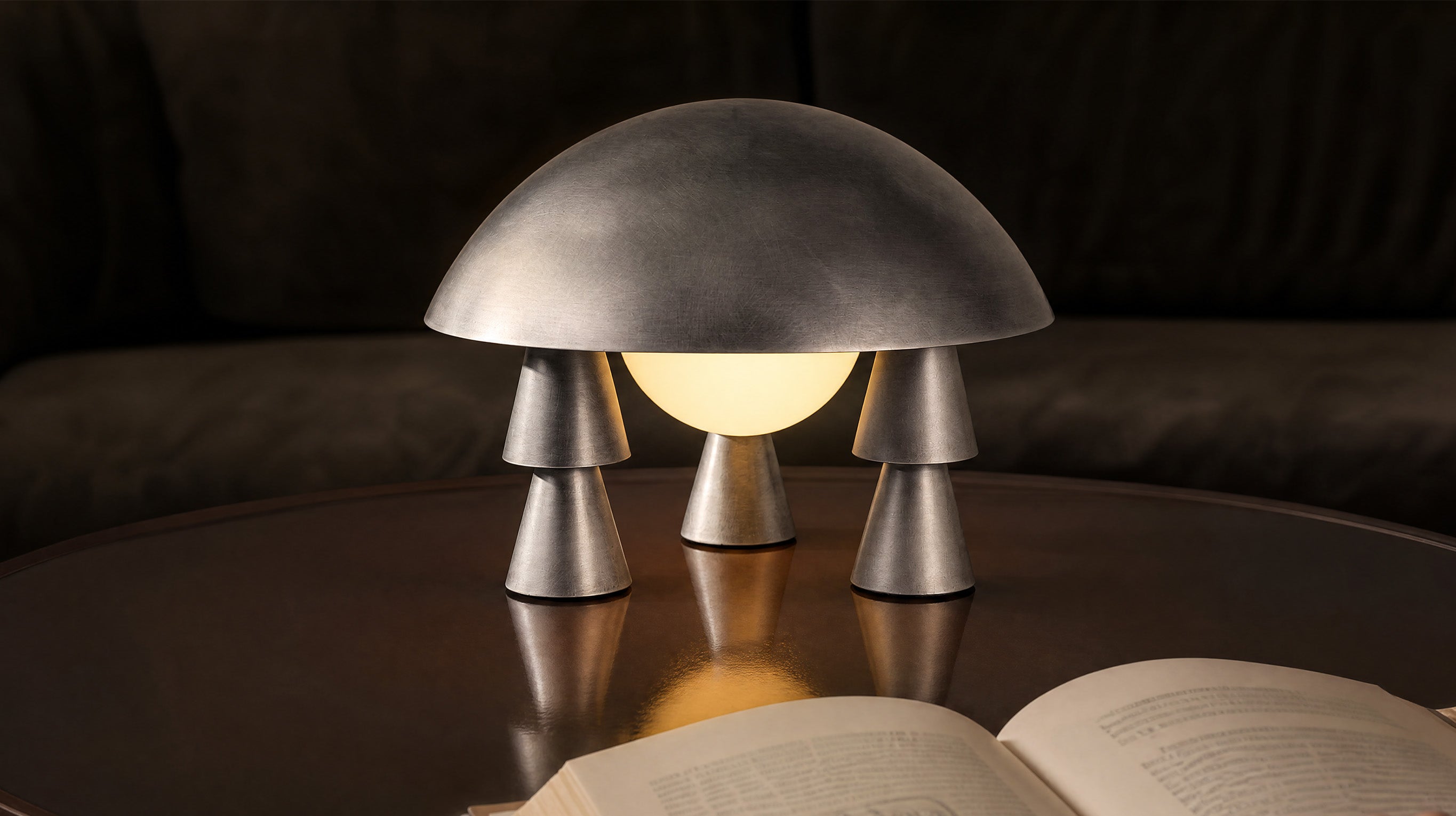 The artamore sculptural metal table lamp features a space-age aesthetic with an aged aluminium finish, emitting a soft ambient glow in a warm minimalist setting.