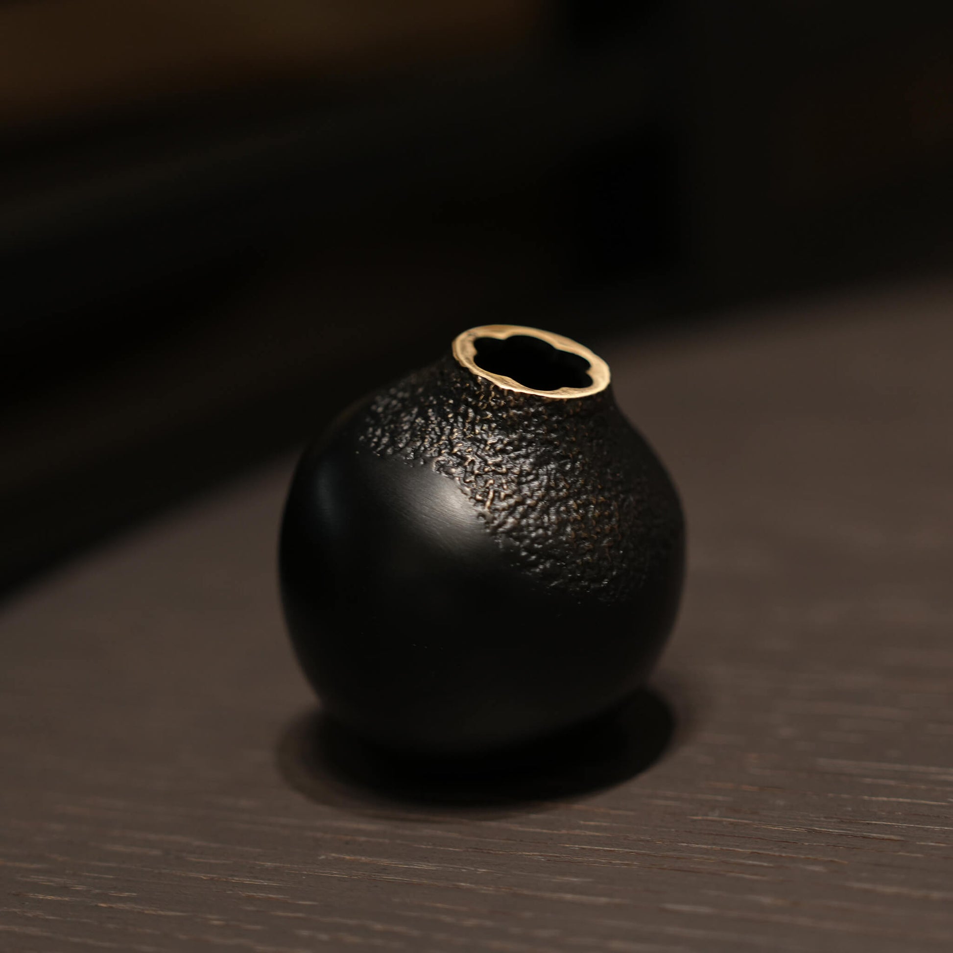 The black textured metal acorn vase placed elegantly on a dark wood table under warm mood lighting, adding wabi-sabi charm