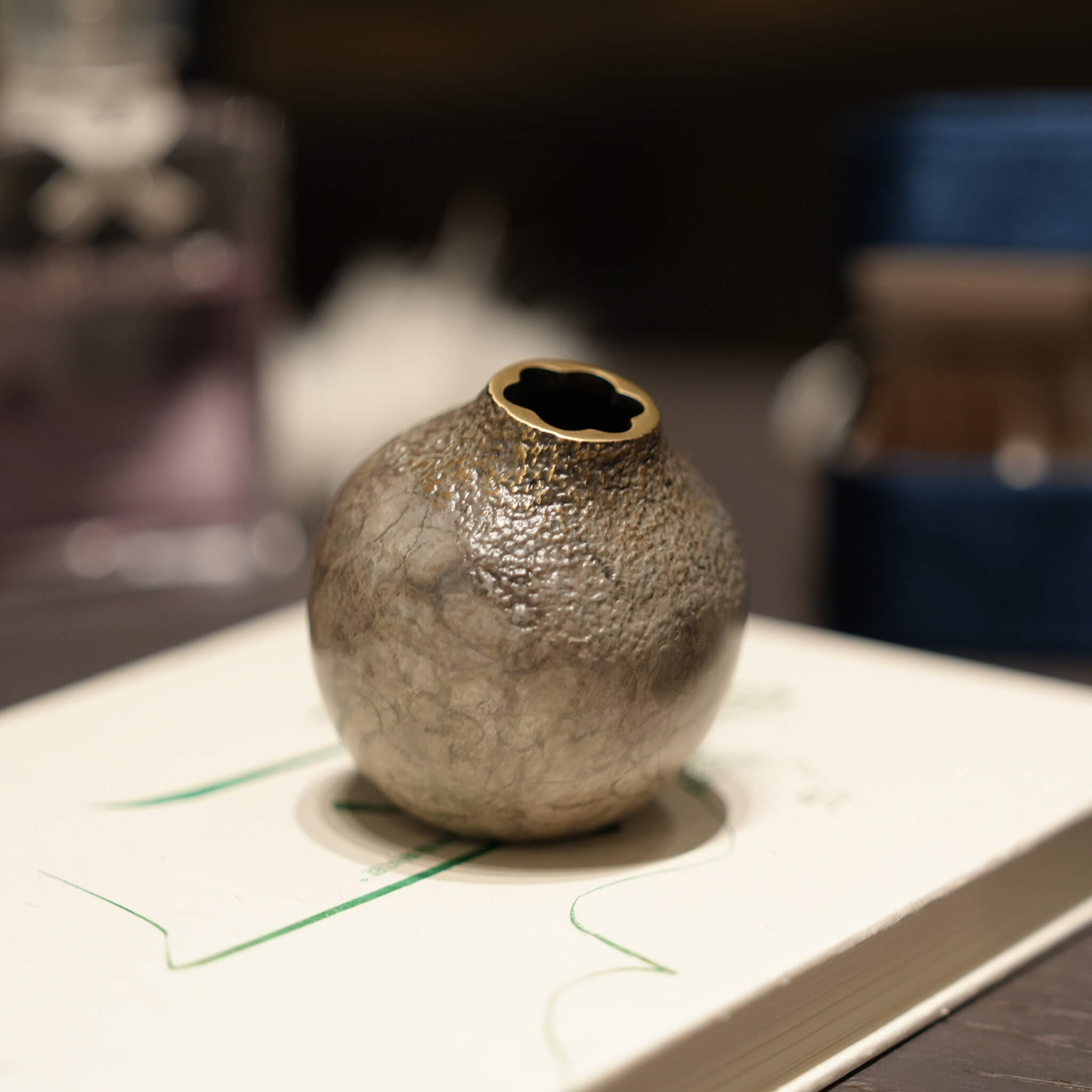 The grey solid brass acorn vase resting on a book in a softly lit, minimalist room, demonstrating its versatile desktop styling