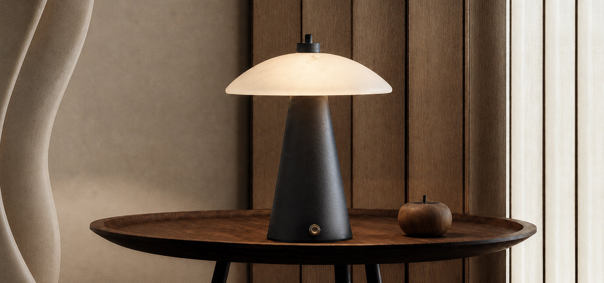 Warm-glowing modern table lamp with a cream dome shade and black tapered base on a dark wooden table.