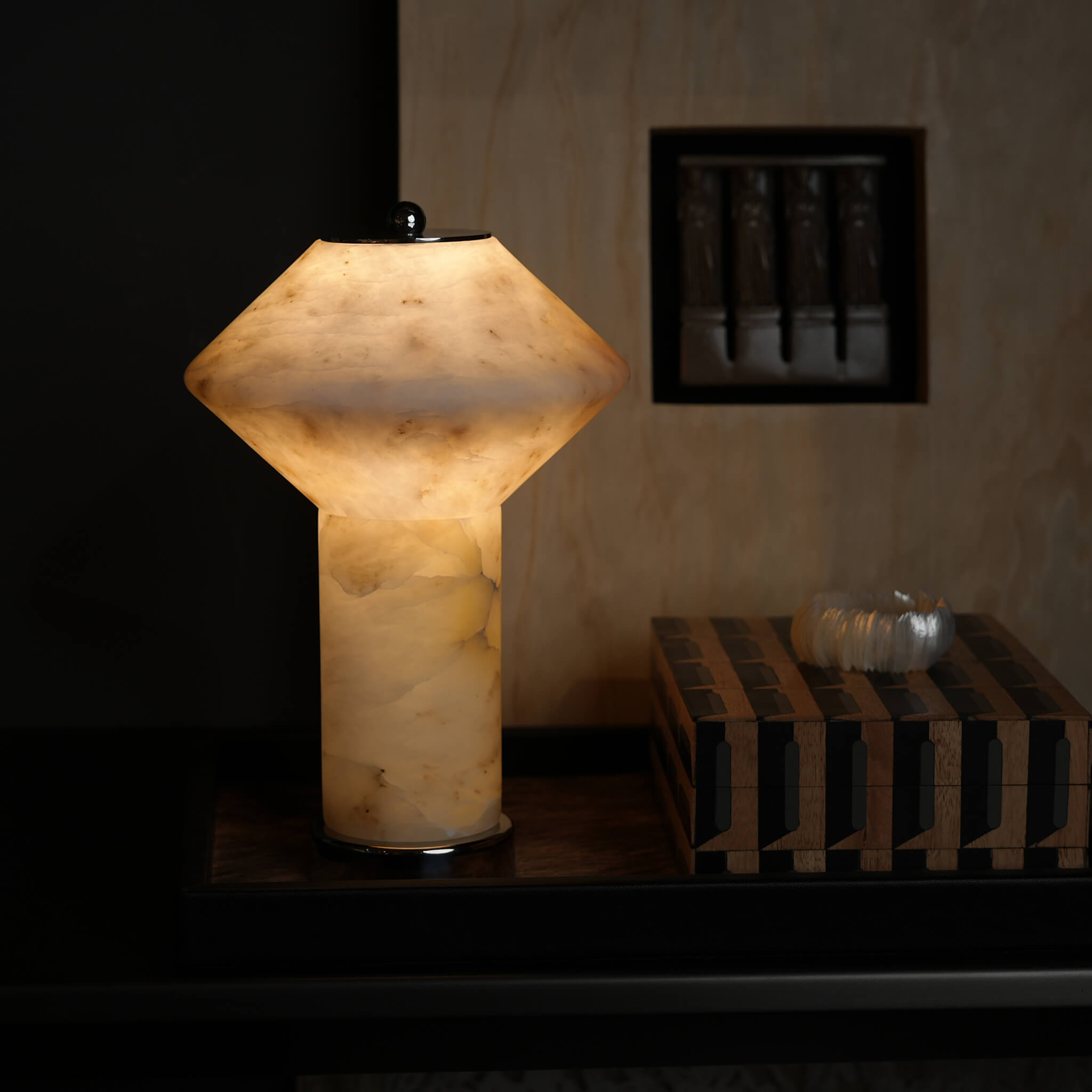 Translucent marble table lamp lit up in a sophisticated, moody study setting next to geometric wooden boxes.