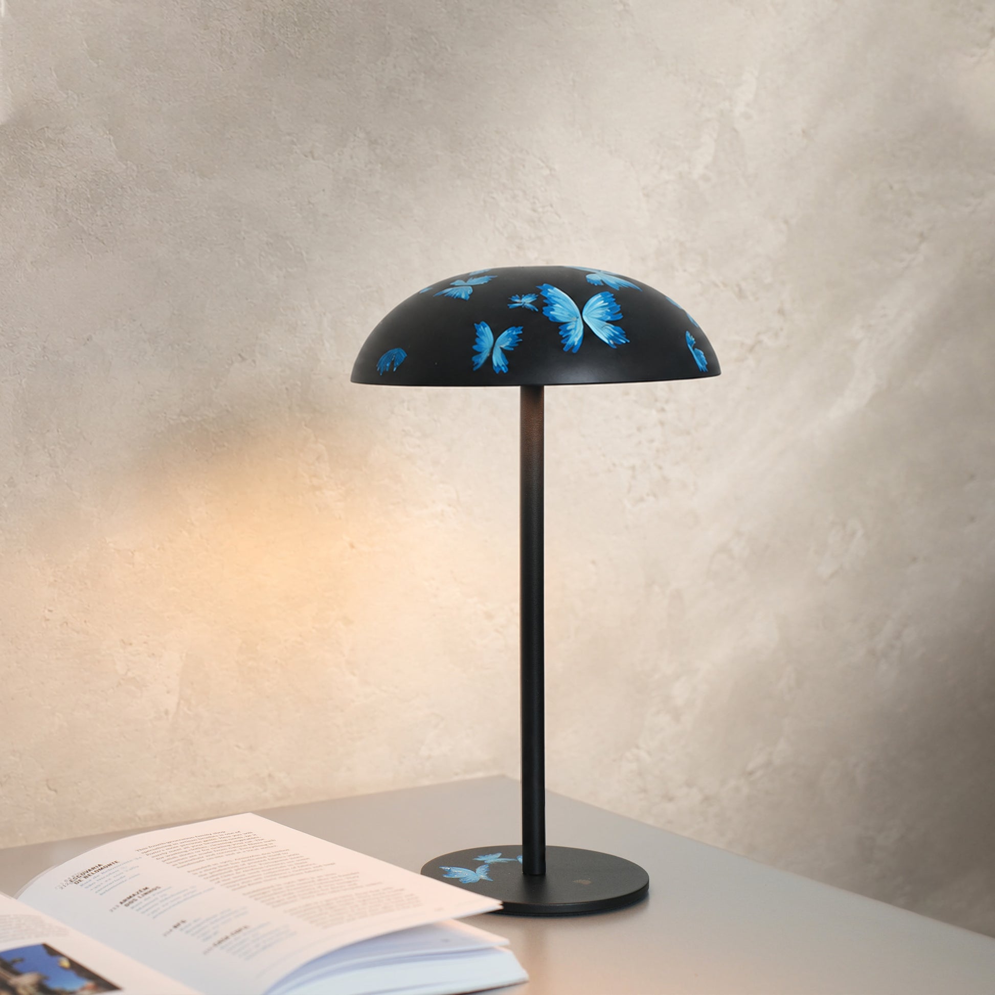 Black Relax-W portable DIY table lamp hand-painted with blue butterflies, glowing beside an open book in a modern interior.