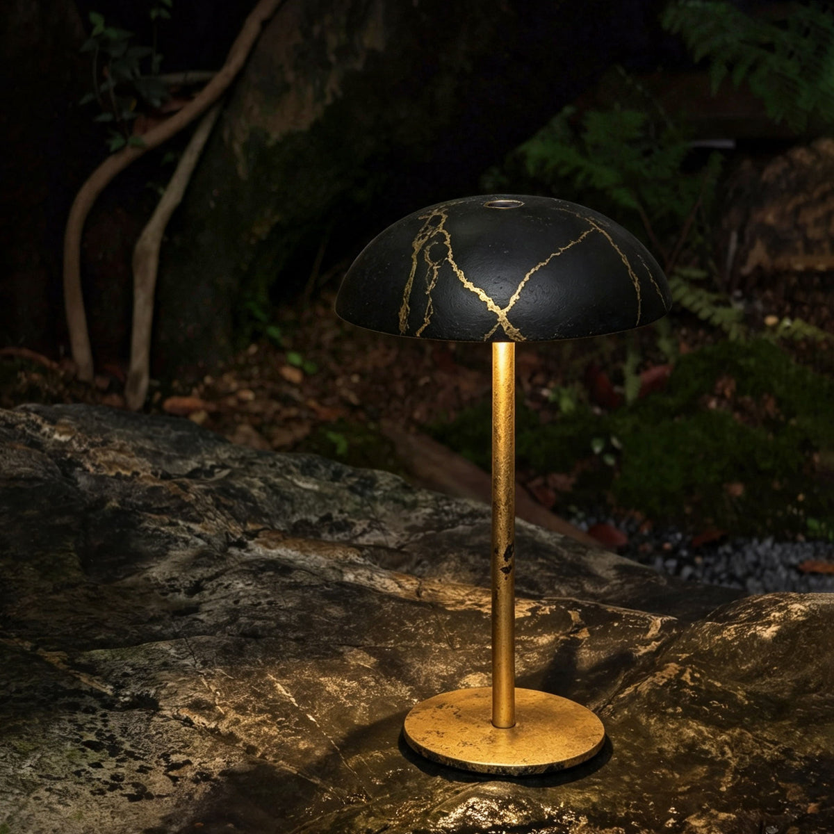 Black and gold hand-painted Relax-W portable table lamp glowing outdoors in a peaceful garden setting.