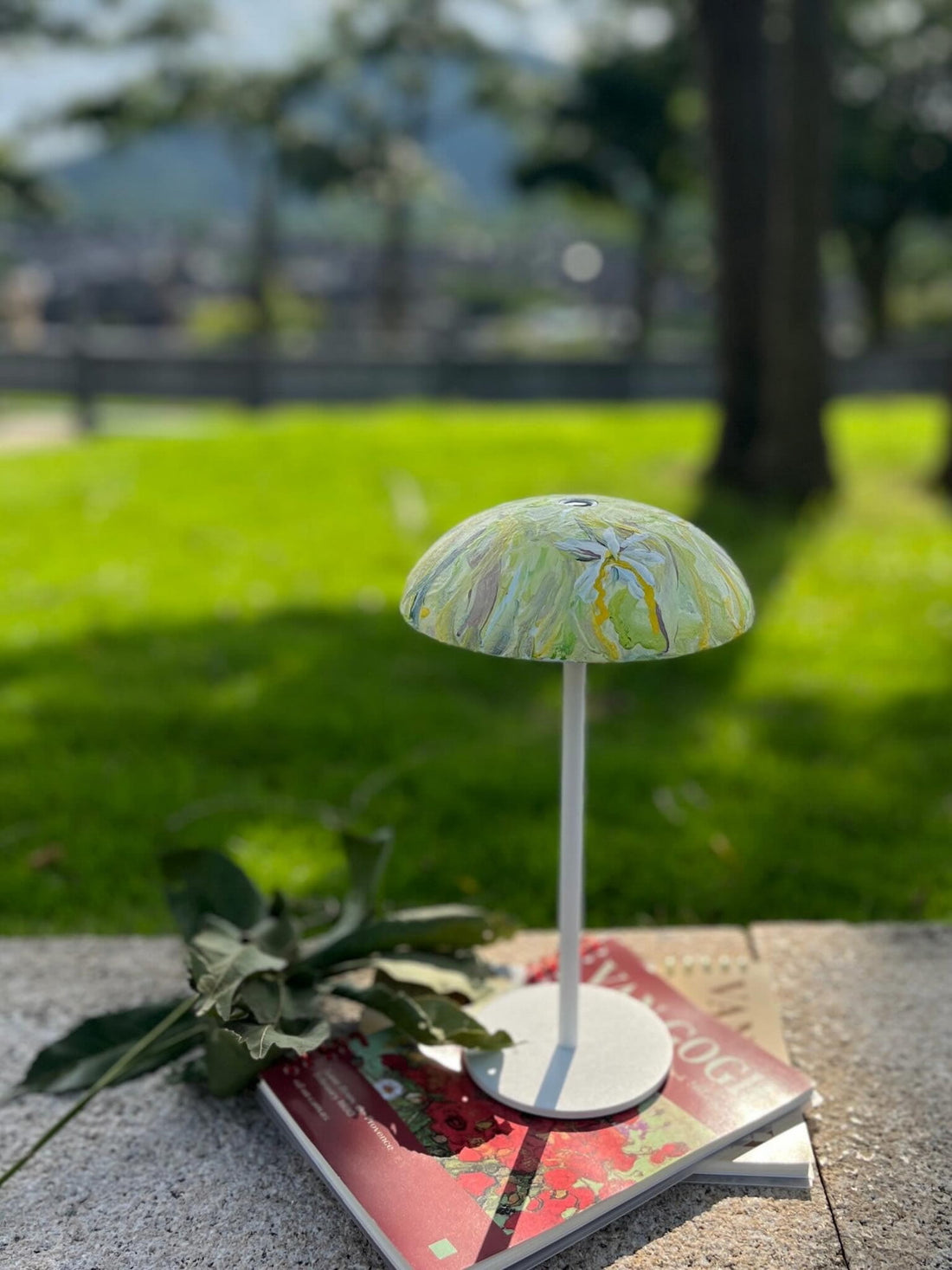 Green floral hand-painted Relax-W DIY portable table lamp displayed outdoors on art books in a sunny garden.