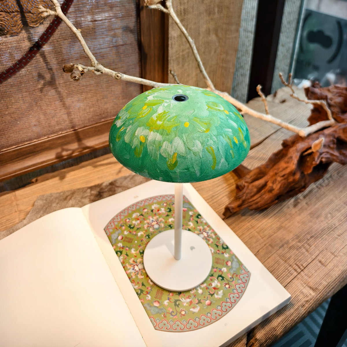 Green floral hand-painted Relax-W DIY table lamp with a white base displayed on an open art book and wooden table.