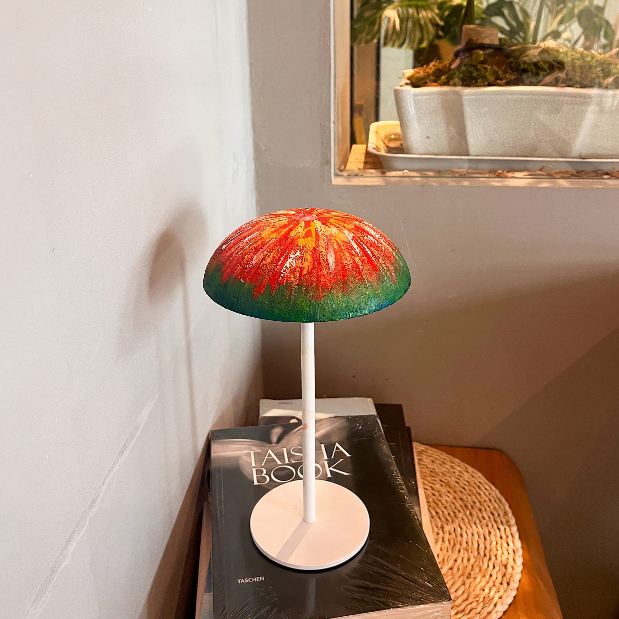 Red and green hand-painted Relax-W portable DIY table lamp with a white base styled on books beside a window.