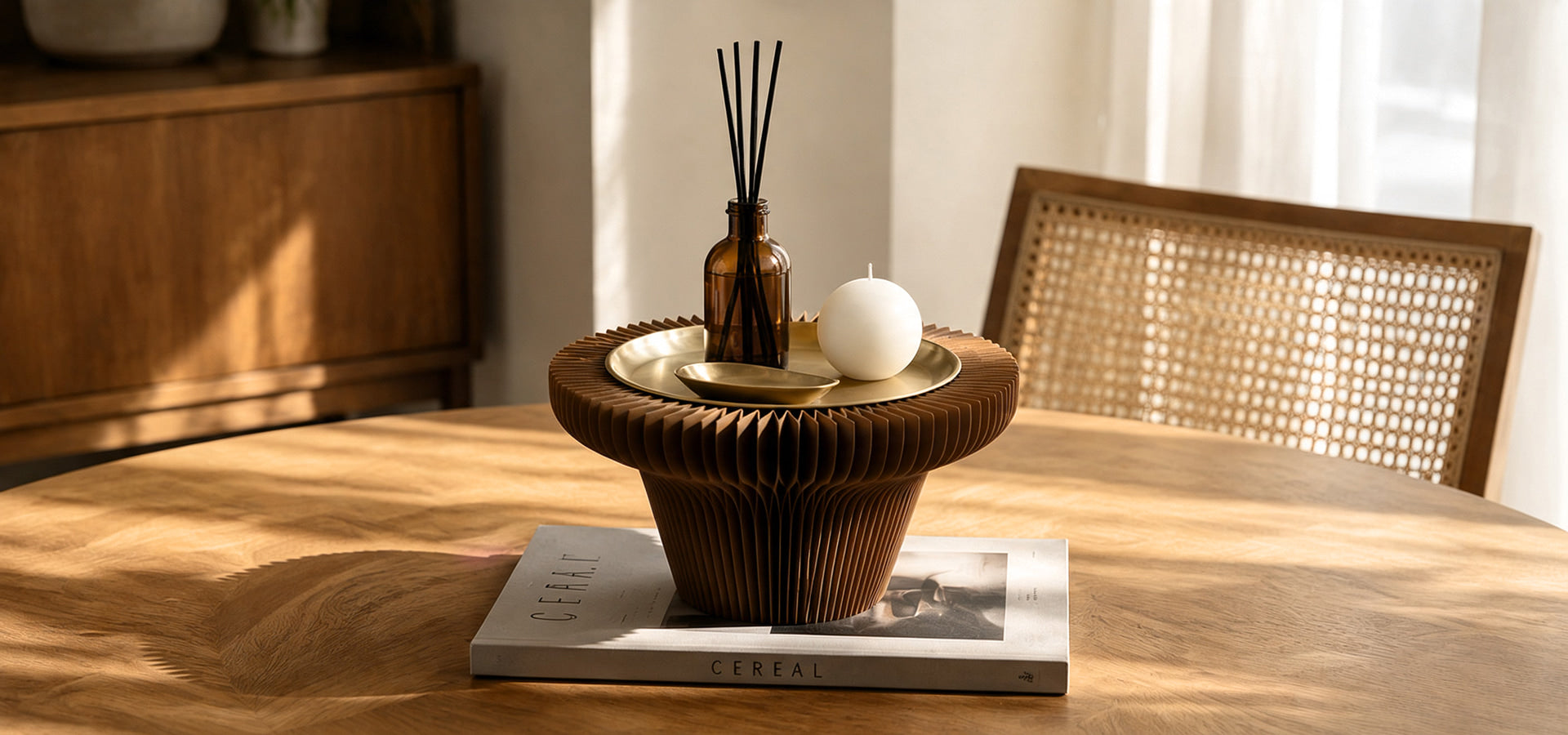 Sculptural wooden table centerpiece with an amber reed diffuser and white candle on a sunlit dining table.