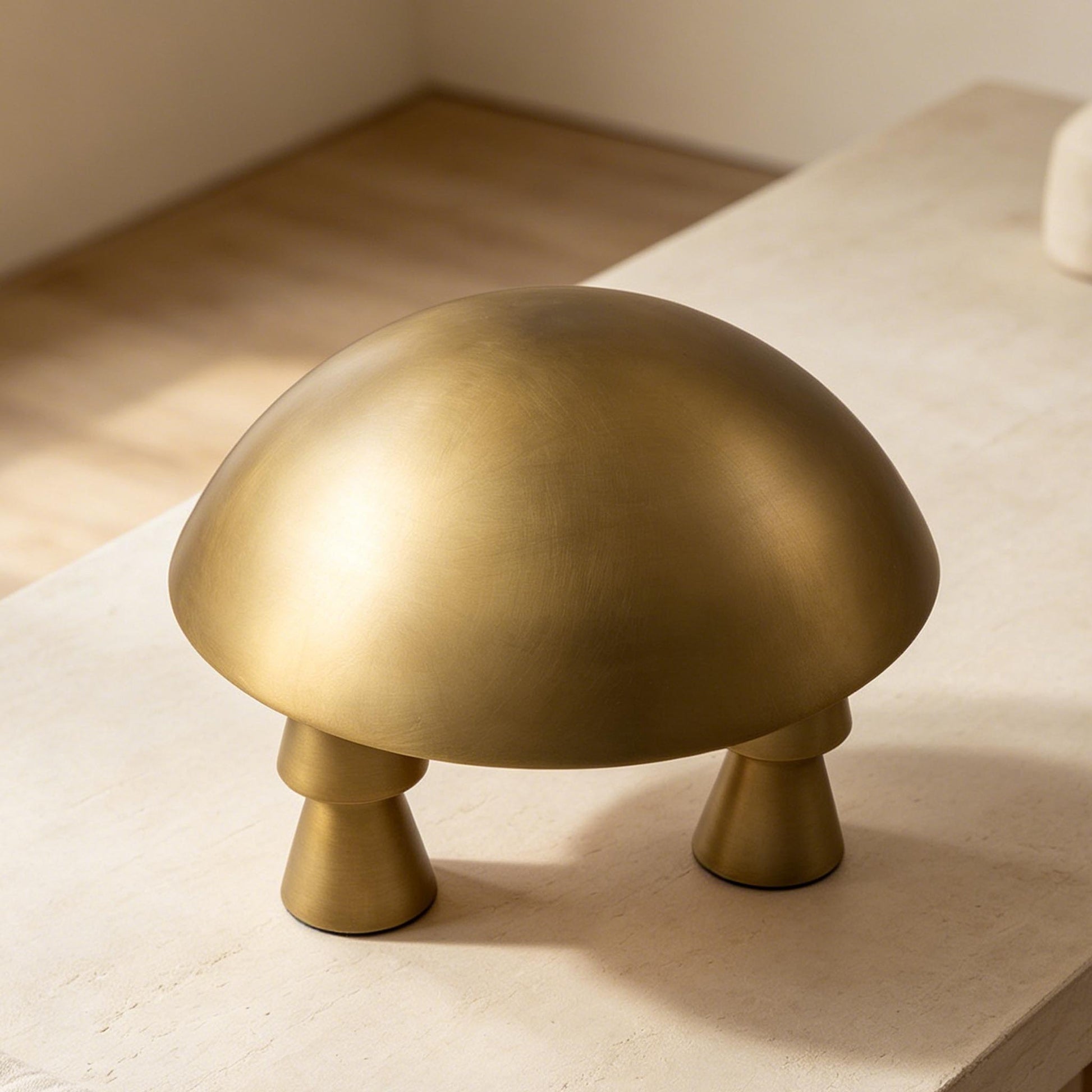 Gold metal table lamp with a smooth dome shade and three conical legs on a light stone tabletop.