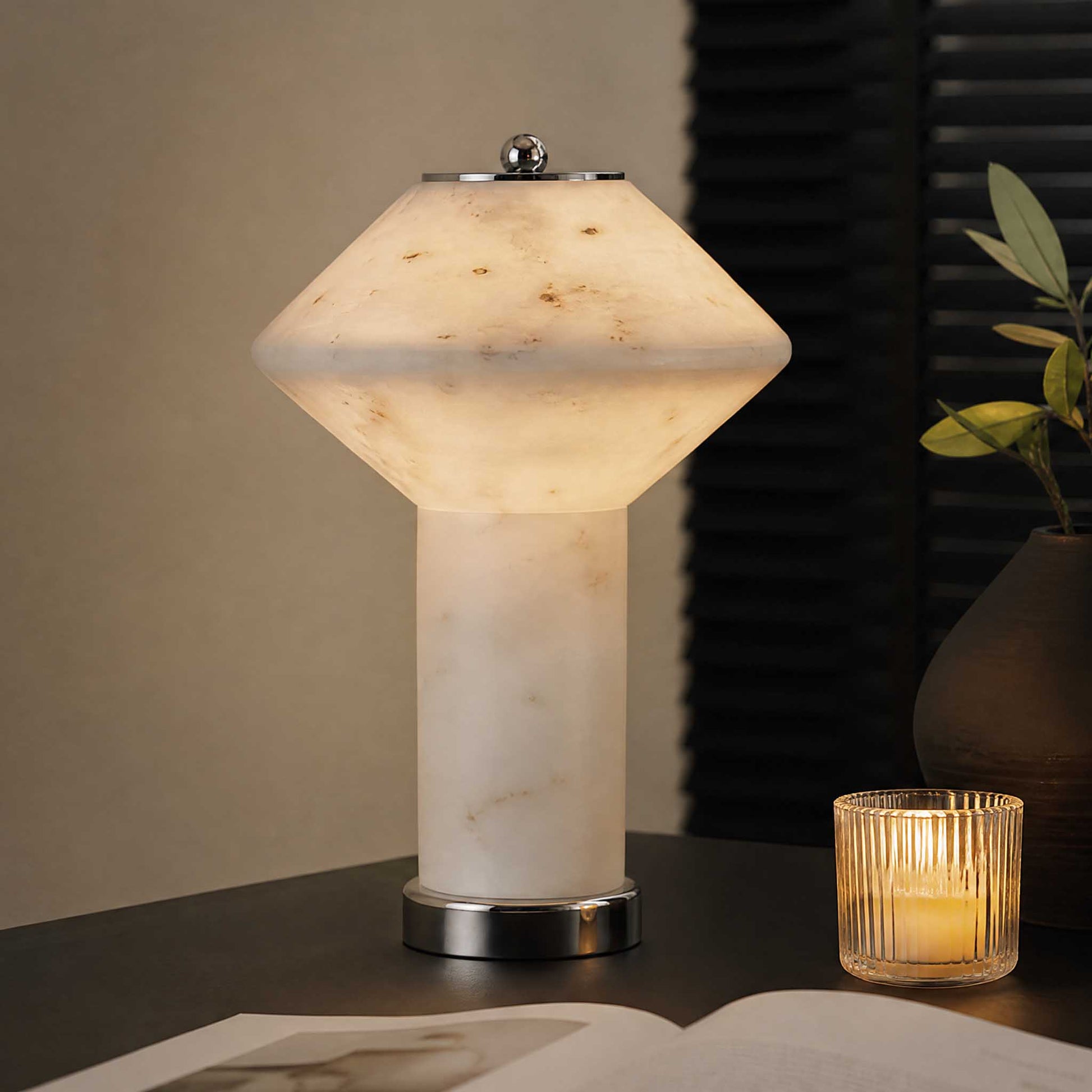 Natural marble table lamp with soft warm light on a dark side table beside a candle and plant.